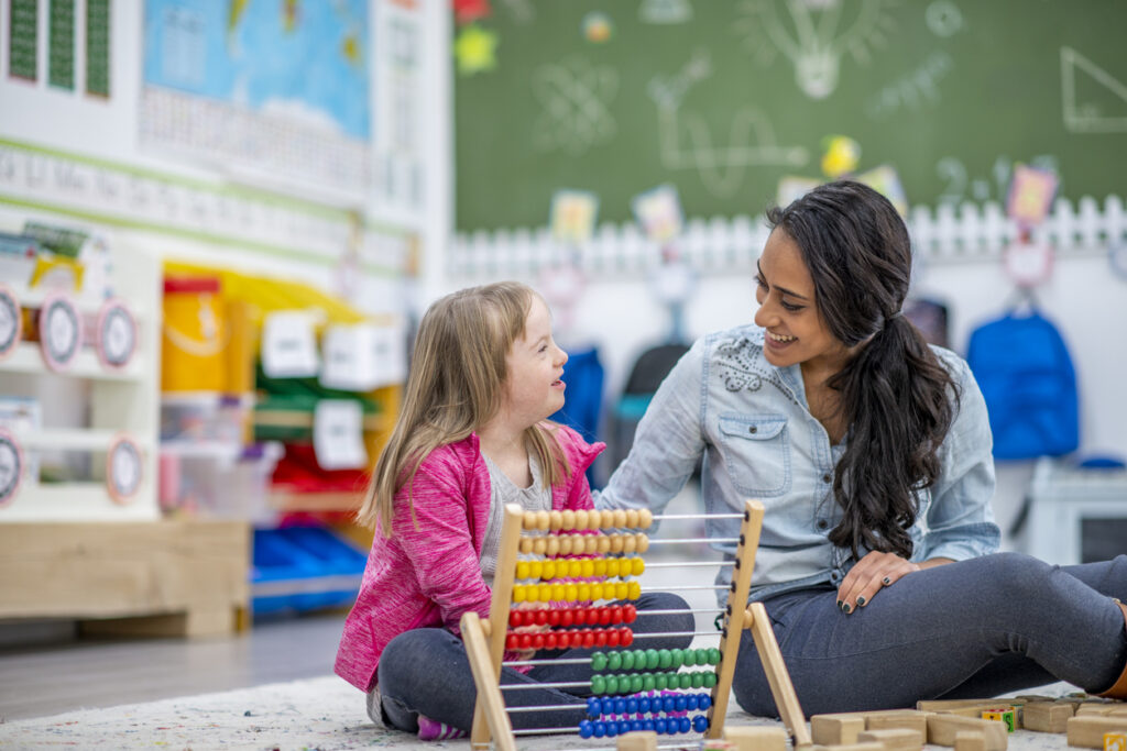 A girl with Down syndrome and her teacher are sitting on the floor in their classroom. The teacher is showing the girl how to use an abacus.